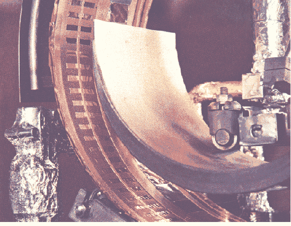 Vista di dettaglio del crogiolo, dello schermo raffreddato, del tamburo con maschere in CuBe su nastro di resina poliimmidica(Kapton). A sinistra il passante a vuoto girevole.